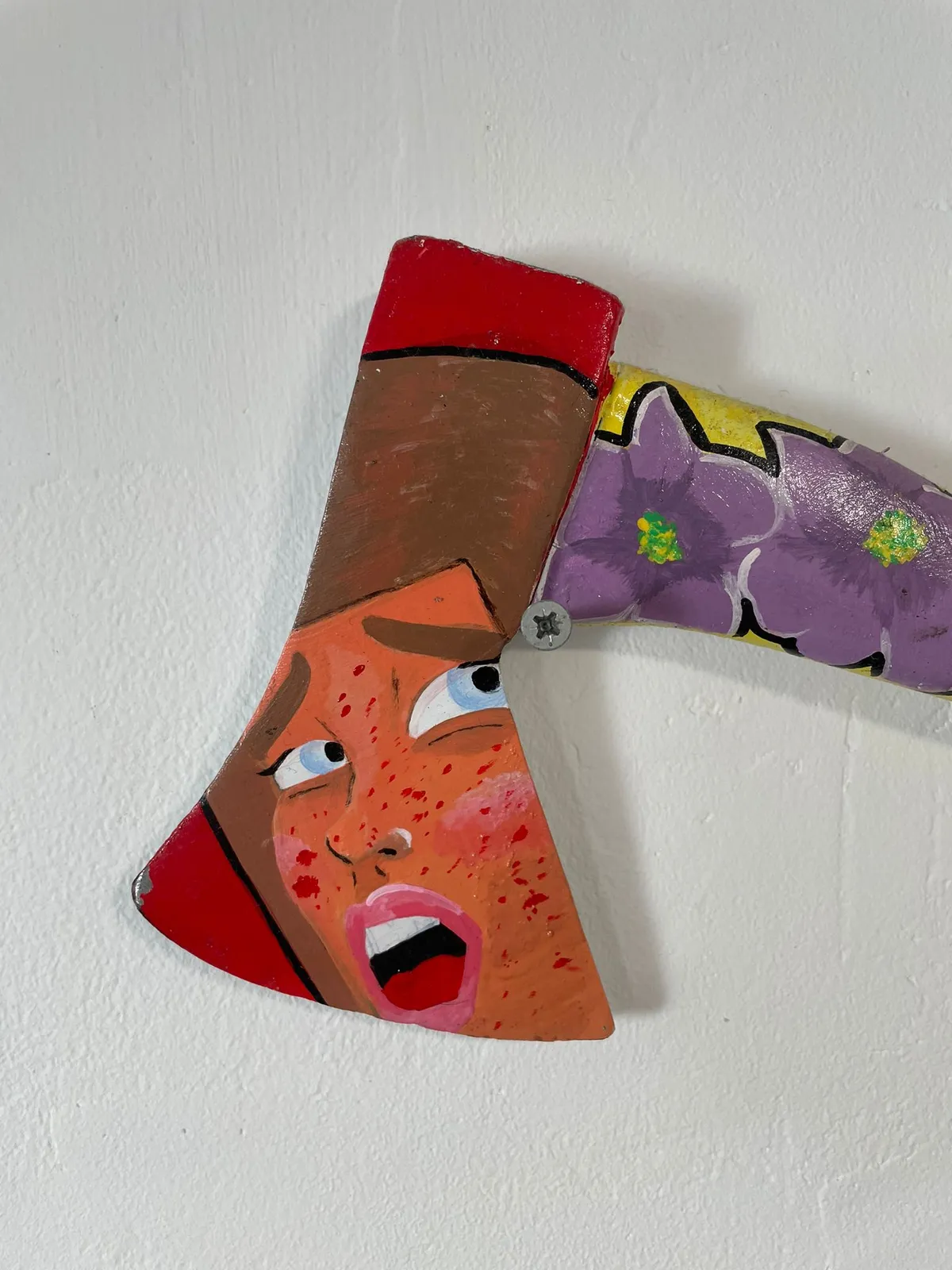 The hatchet mounted on a white wall, seen at an angle so the painted face on the blade catches the light and the purple flowers run away down the handle.