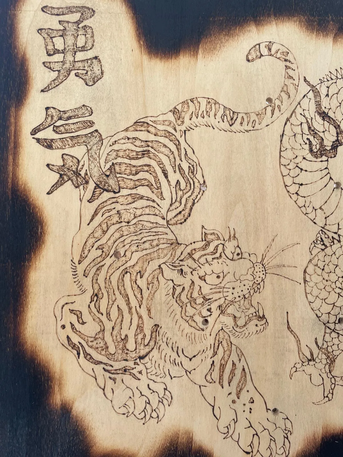 Detail of the burned tiger prowling beneath two kanji characters, its stripes and face built from short burned strokes against the charred edge of the panel.